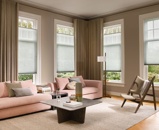 Woonkamer met lichte plissé raamdecoratie, beige gordijnen, roze bank en houten stoel