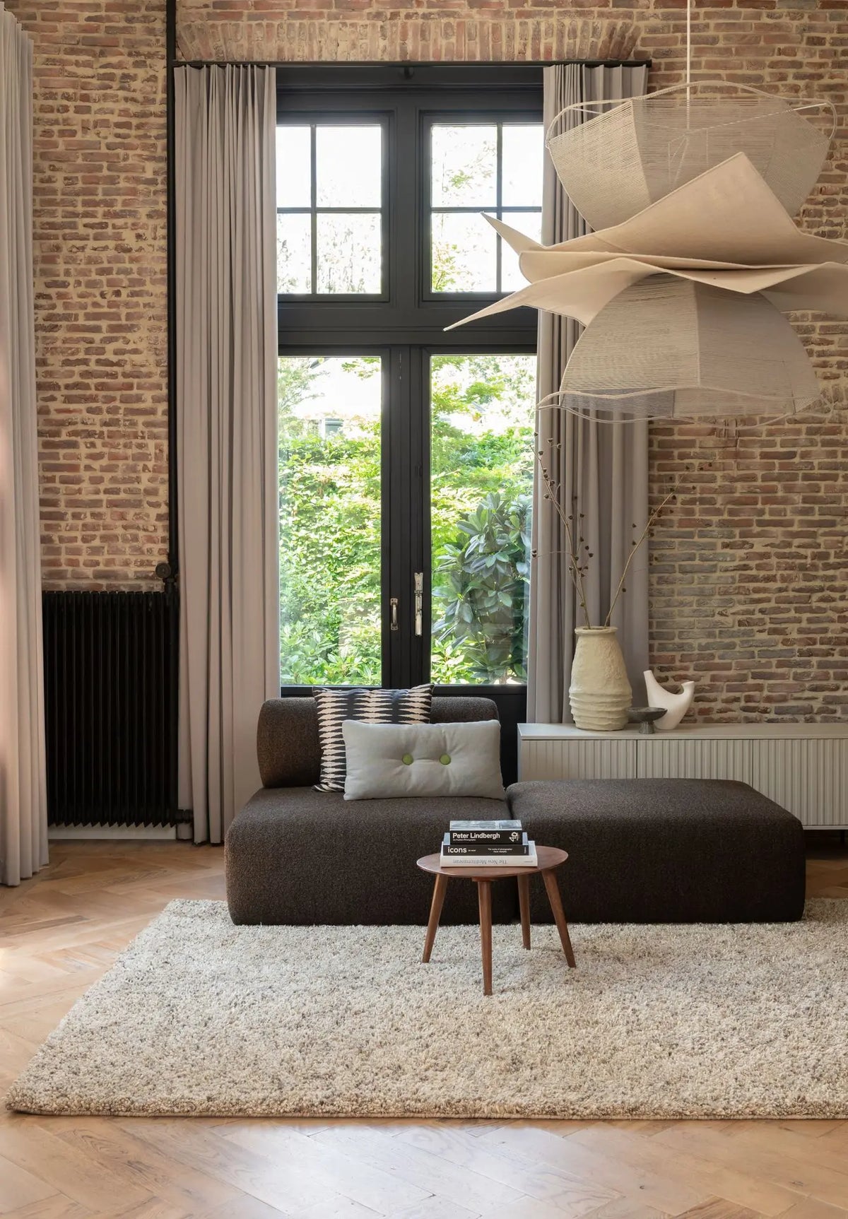 Woonkamer met naturel hoogpolig vloerkleed, bruine bank, gordijnen en bakstenen wand