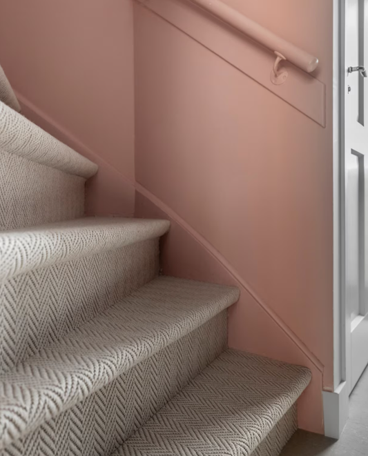 Trap met grijs-wit visgraat tapijt als trapbekleding langs een roze wand