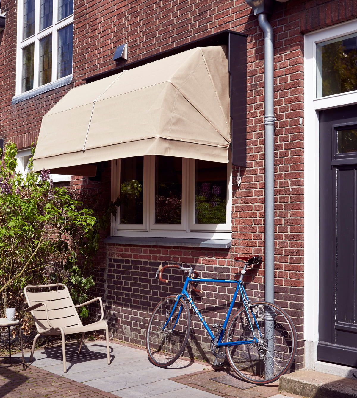 Bakstenen gevel met crèmekleurige korfmarkies boven het raam, tuinstoel en blauwe fiets
