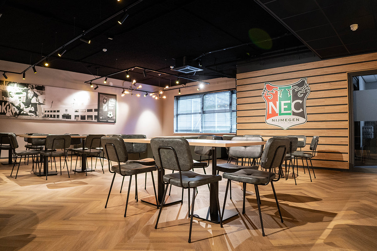 Vernieuwde spelershome van NEC Nijmegen met visgraatvloer, tafels, stoelen en NEC-logo op houten wand