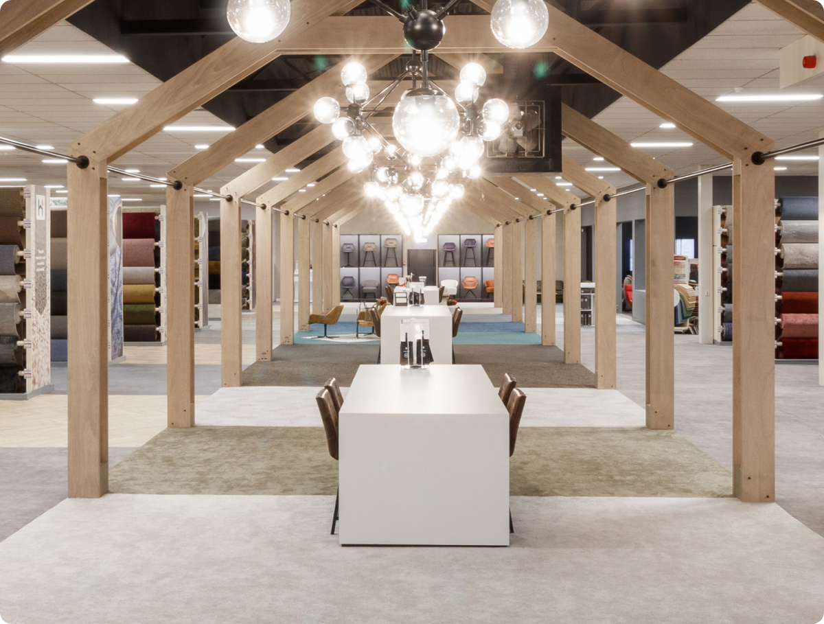 Showroom van Hofmans at Home met houten dakspanten, kroonluchters en lange tafel tussen tapijtrollen
