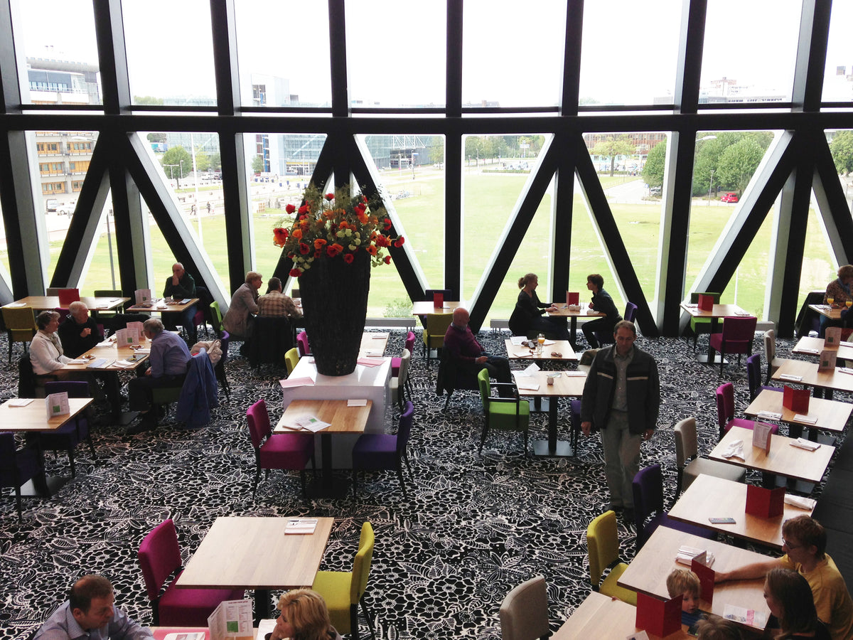 Restaurant met grote glazen gevel, gedekte tafels, kleurrijke stoelen en zwart-wit tapijt