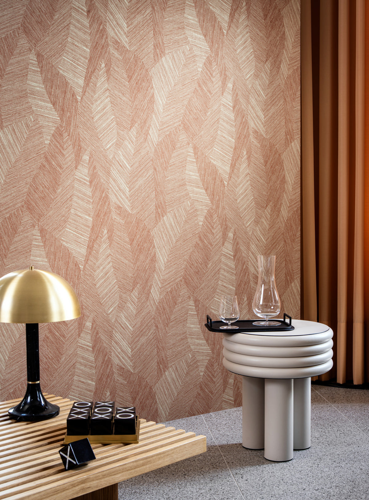 Modern interieur met terra-roze behang in geometrisch bladmotief, gecombineerd met warme gordijnen, een gouden tafellamp en een rond bijzettafeltje met glazen karaf en glazen.