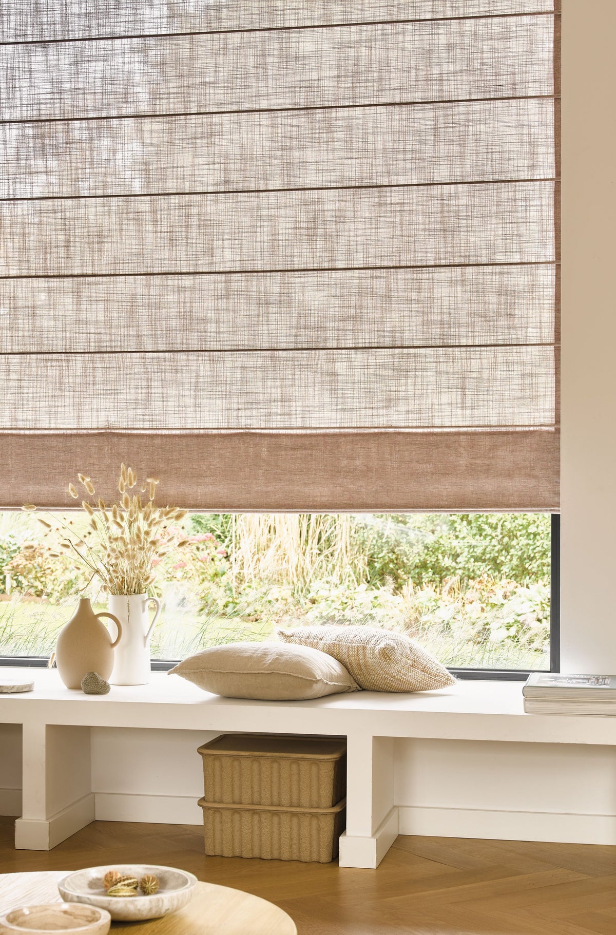 Houten vouwgordijn in natuurlijke beige tint, met een lichte doorkijk, boven een zithoek met kussens en decoratieve vazen in een warme, serene woonkamer.