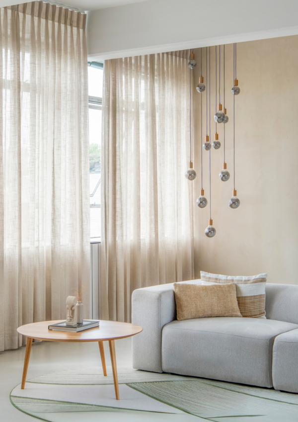 Lichte zithoek met grijze bank, ronde salontafel en beige vitragegordijnen voor hoge ramen