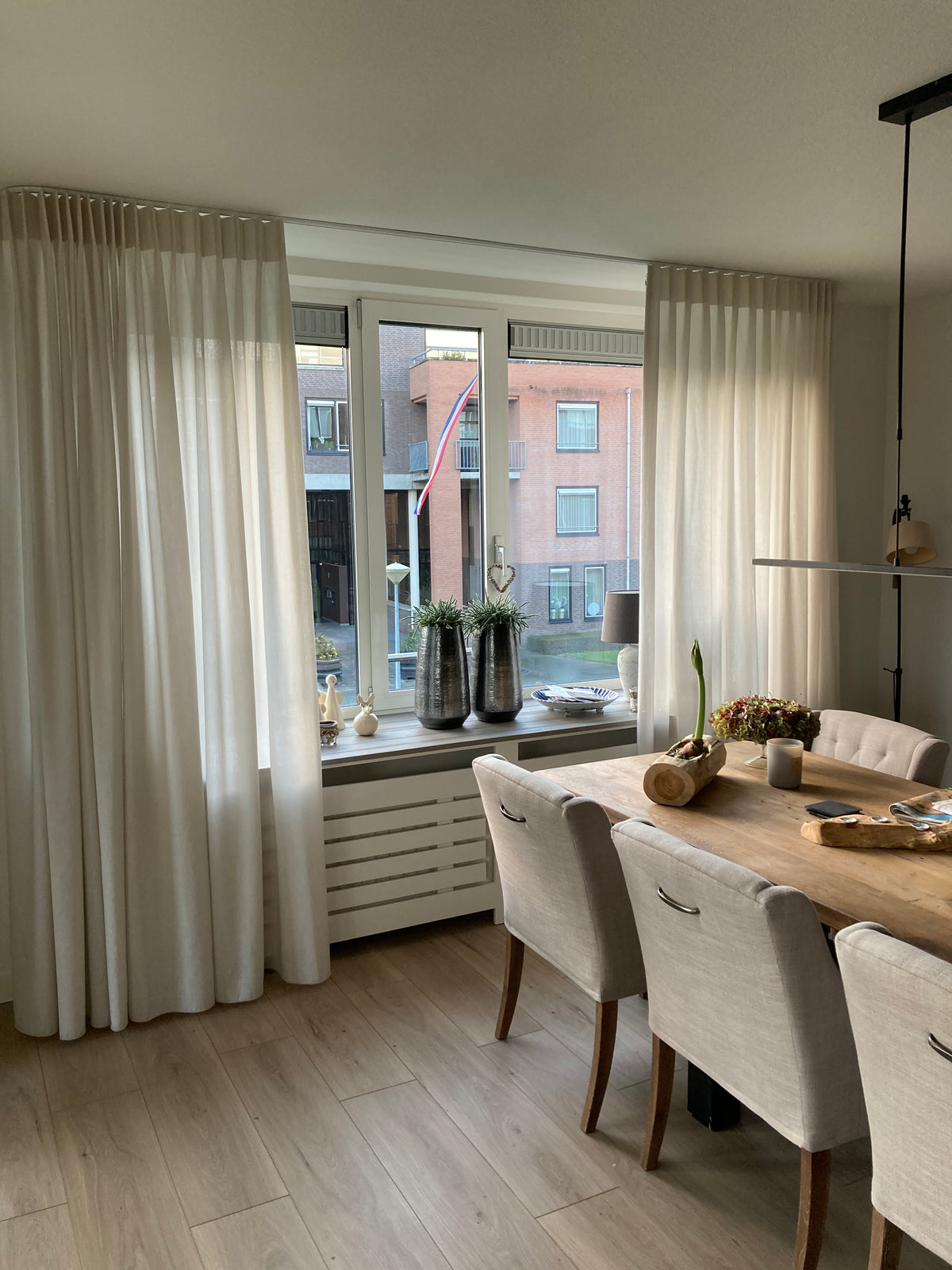 Eetkamer met lange houten tafel, beige stoelen, witte gordijnen en lichte houtlook vloer