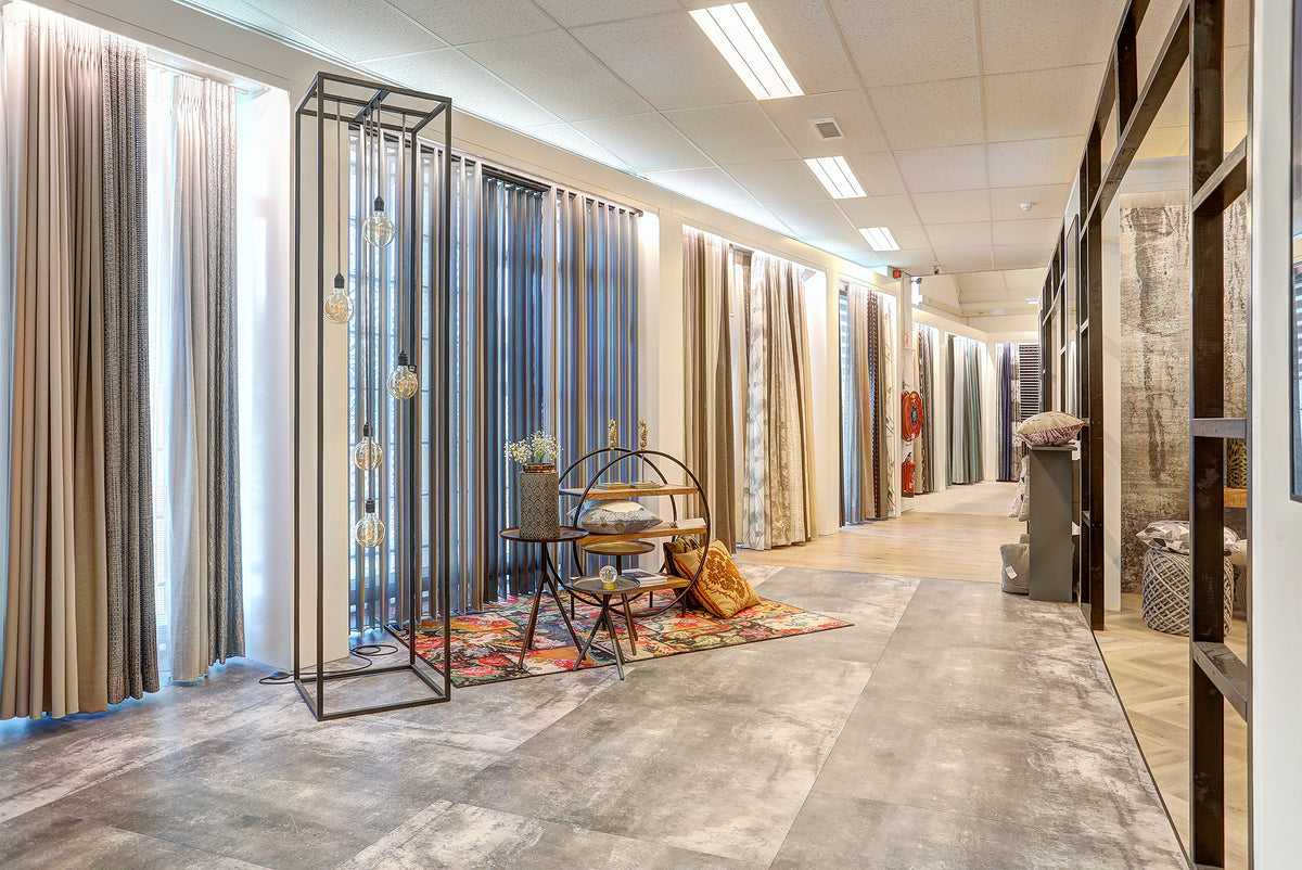 Showroompresentatie van gordijnen en raamdecoratie bij Hofmans, vitrage en inbetweens met diverse stoffen, kleuren en structuren naast een betonlook PVC-vloer en sfeervolle styling