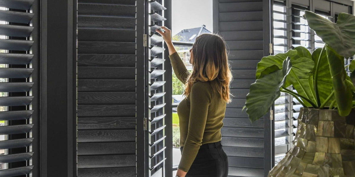 Vrouw opent zwarte houten shutters voor een raam, naast een grote kamerplant
